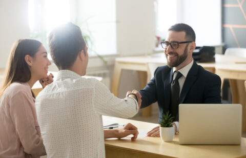 A smiling financial advisor in a suit shakes hands with a male client, while a female client watches with a smile in a modern office setting.