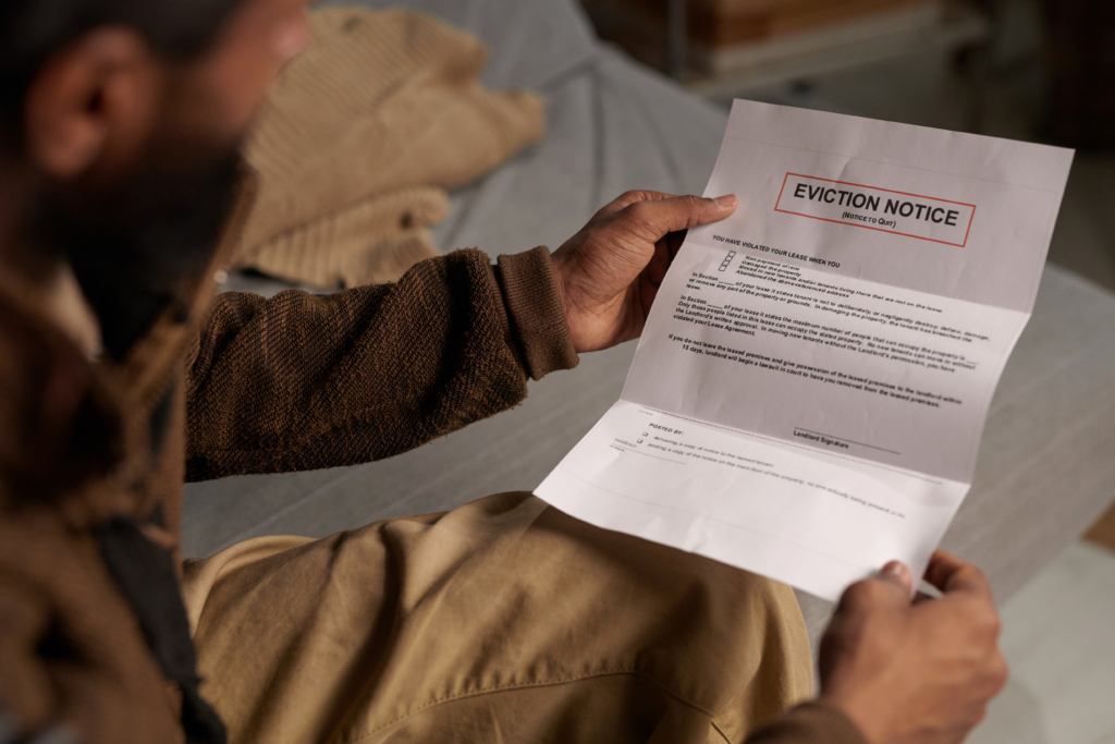 A person wearing a brown sweater and beige pants sits on a bed, holding and reading an eviction notice with a serious expression.