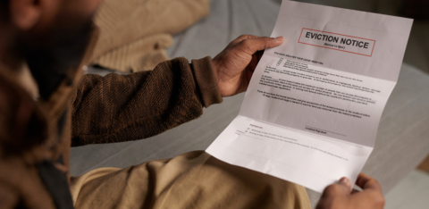 A person wearing a brown sweater and beige pants sits on a bed, holding and reading an eviction notice with a serious expression.
