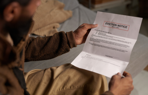 A person wearing a brown sweater and beige pants sits on a bed, holding and reading an eviction notice with a serious expression.
