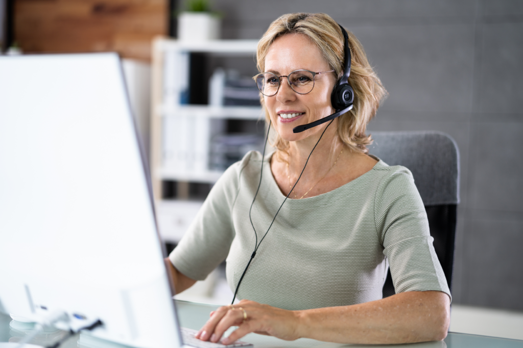 A smiling middle-aged woman wearing a headset works on her computer in a modern office setting.