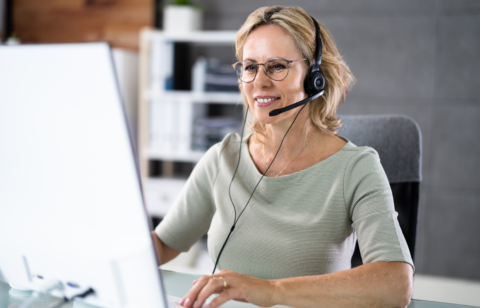 A smiling middle-aged woman wearing a headset works on her computer in a modern office setting.