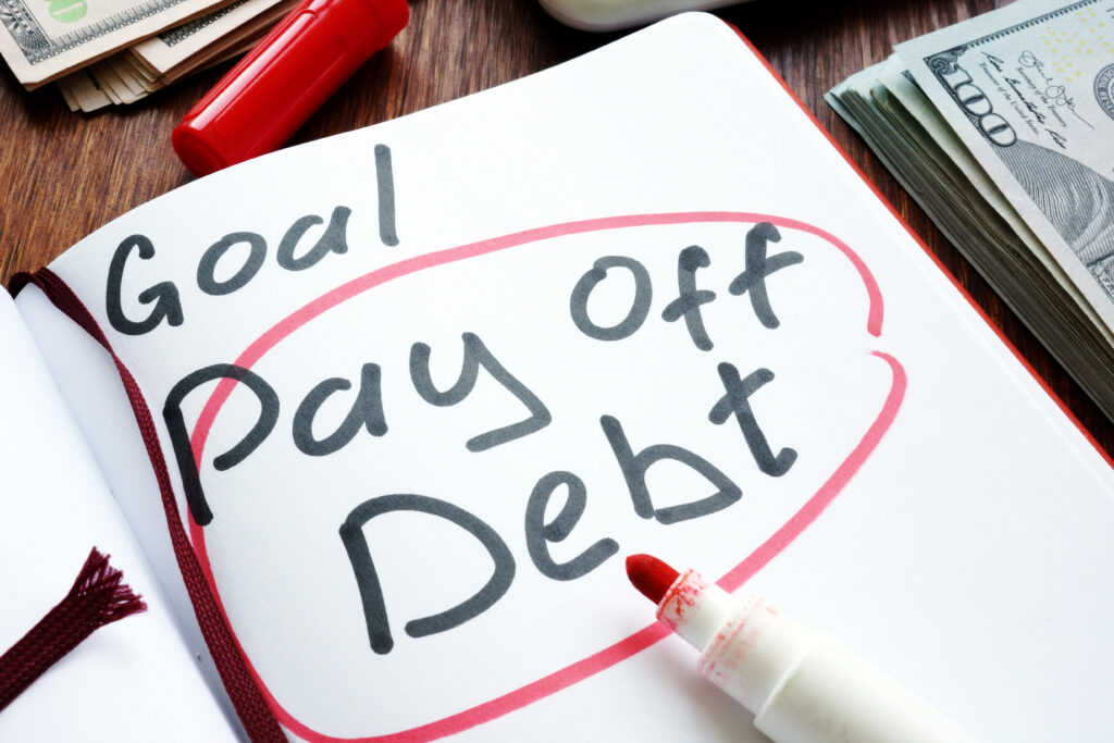 Notebook with "Goal: Pay Off Debt" written in bold letters, surrounded by a red circle, next to a red marker and stacks of cash.