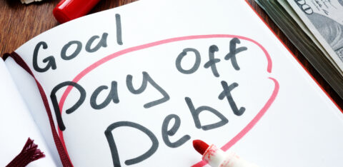 Notebook with "Goal: Pay Off Debt" written in bold letters, surrounded by a red circle, next to a red marker and stacks of cash.