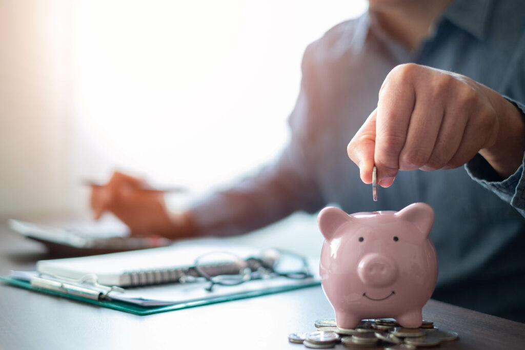 A person placing a coin into a pink piggy bank while working on financial planning with a calculator and documents in the background.