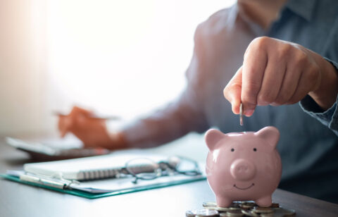 A person placing a coin into a pink piggy bank while working on financial planning with a calculator and documents in the background.