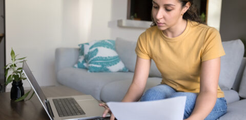 Young woman sitting on a couch reviewing financial documents with a calculator and laptop on the table.