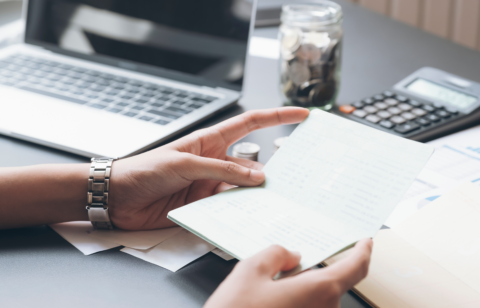Hands holding a bank statement or financial document at a desk with a laptop, calculator, and jar of coins in the background.