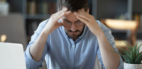 Stressed man sitting at a desk with his hands on his head, looking at financial documents and a calculator.