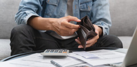 Man sitting on a couch checking his empty wallet, surrounded by bills, a calculator, and a laptop.