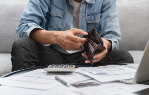Man sitting on a couch checking his empty wallet, surrounded by bills, a calculator, and a laptop.