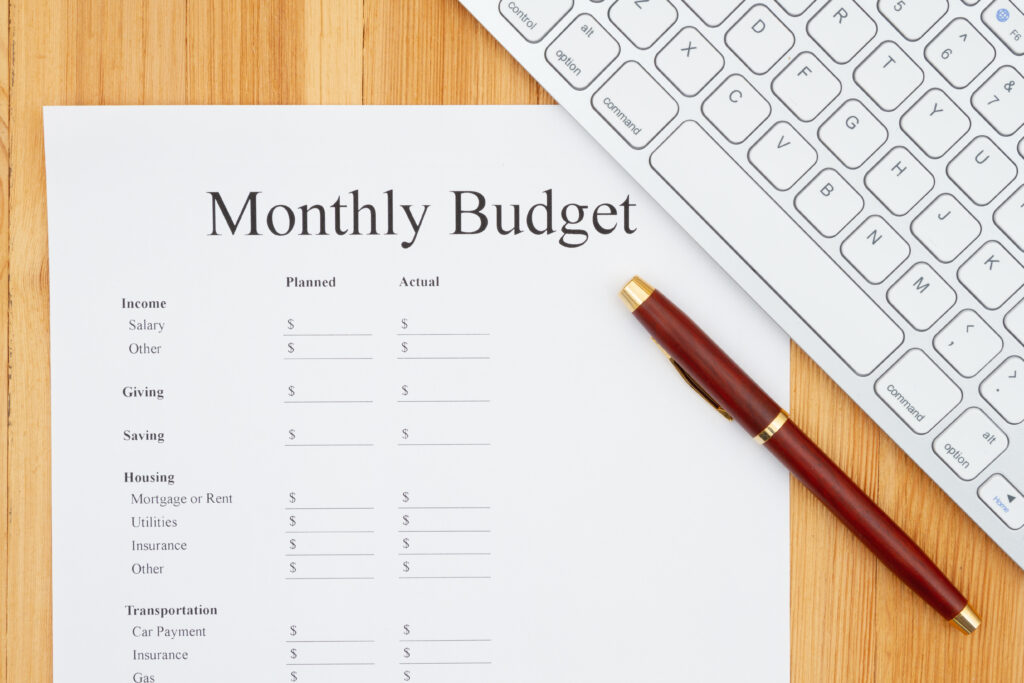 A "Monthly Budget" worksheet on a wooden desk, accompanied by a red and gold pen and a partial view of a white computer keyboard.