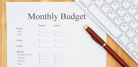 A "Monthly Budget" worksheet on a wooden desk, accompanied by a red and gold pen and a partial view of a white computer keyboard.