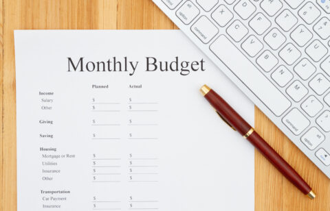 A "Monthly Budget" worksheet on a wooden desk, accompanied by a red and gold pen and a partial view of a white computer keyboard.