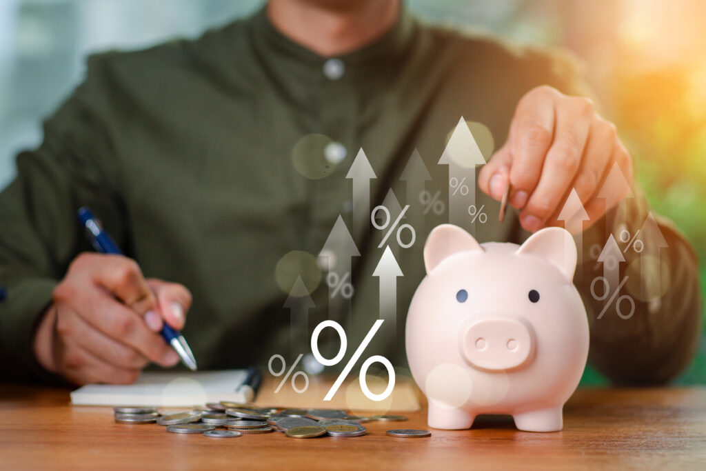 A person placing a coin into a piggy bank while writing in a notebook, with digital percentage symbols and upward arrows representing growing savings.
