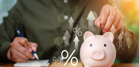 A person placing a coin into a piggy bank while writing in a notebook, with digital percentage symbols and upward arrows representing growing savings.