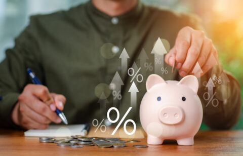 A person placing a coin into a piggy bank while writing in a notebook, with digital percentage symbols and upward arrows representing growing savings.