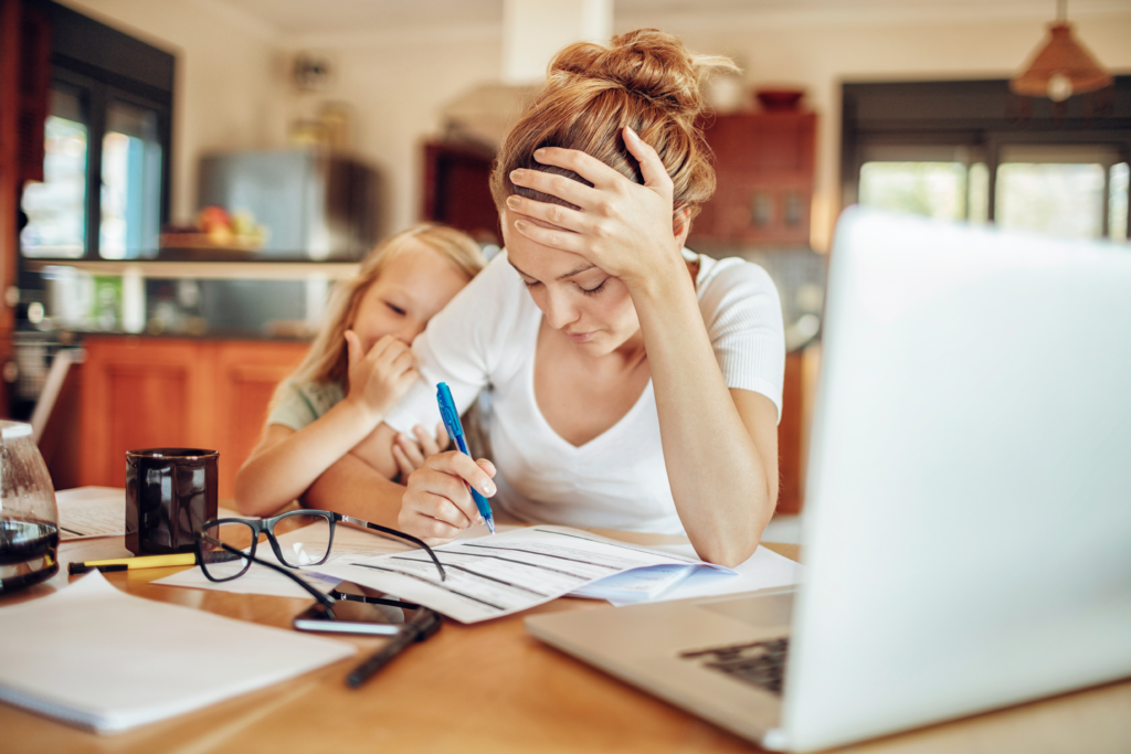 A stressed mother sits at a kitchen table, reviewing financial documents with a pen in hand, while her young daughter leans on her for comfort.
