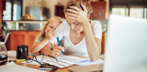 A stressed mother sits at a kitchen table, reviewing financial documents with a pen in hand, while her young daughter leans on her for comfort.