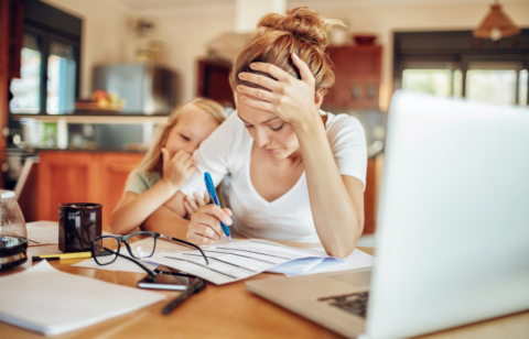 A stressed mother sits at a kitchen table, reviewing financial documents with a pen in hand, while her young daughter leans on her for comfort.