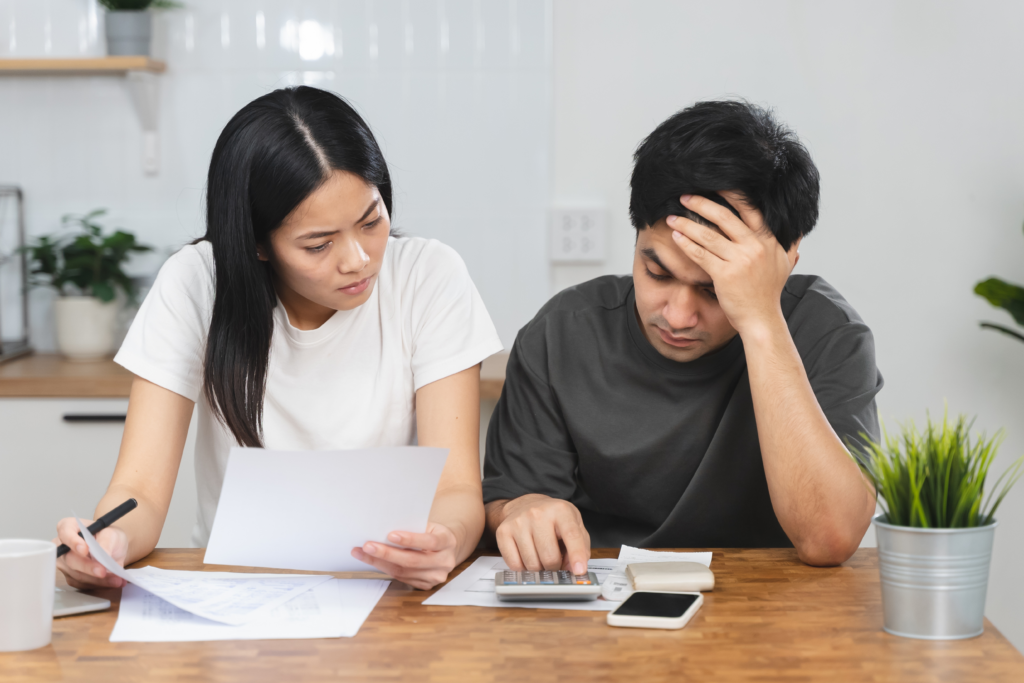 Stressed couple sitting at a table reviewing financial documents and using a calculator to manage expenses.