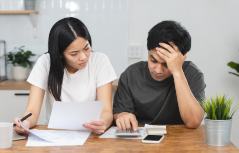 Stressed couple sitting at a table reviewing financial documents and using a calculator to manage expenses.