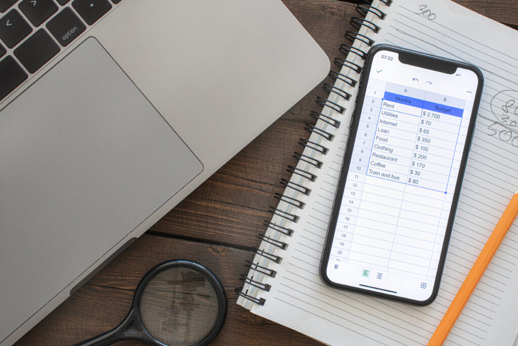 A smartphone displaying a digital budgeting spreadsheet next to a laptop, notebook, and pencil on a wooden desk.