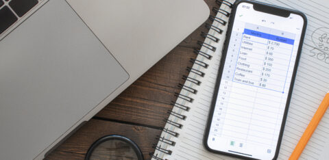 A smartphone displaying a digital budgeting spreadsheet next to a laptop, notebook, and pencil on a wooden desk.