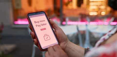 Person holding a smartphone displaying a "Buy now, pay later" message on a shopping app screen, with a blurred outdoor café background.