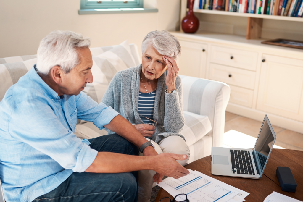 Older couple sitting on a couch reviewing financial documents with a laptop, looking concerned while discussing their finances.