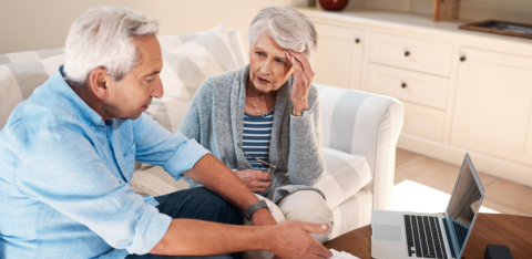 Older couple sitting on a couch reviewing financial documents with a laptop, looking concerned while discussing their finances.