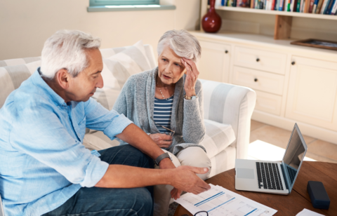Older couple sitting on a couch reviewing financial documents with a laptop, looking concerned while discussing their finances.