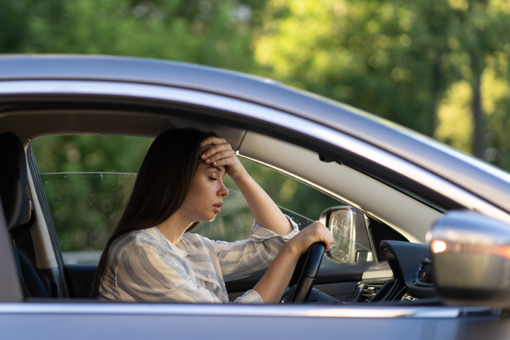 A woman sitting in a car with her hand on her forehead, looking stressed and overwhelmed.