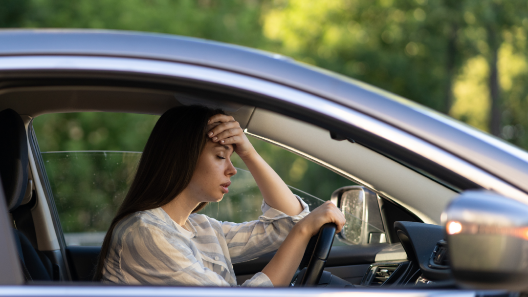 A woman sitting in a car with her hand on her forehead, looking stressed and overwhelmed.