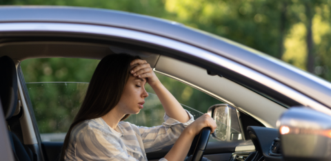 A woman sitting in a car with her hand on her forehead, looking stressed and overwhelmed.