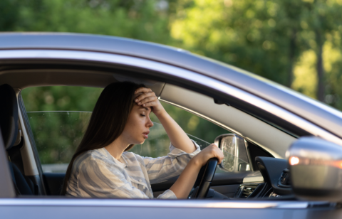 A woman sitting in a car with her hand on her forehead, looking stressed and overwhelmed.