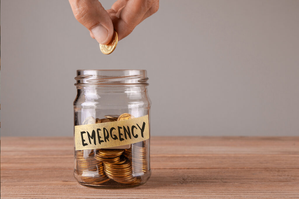 Hand placing a coin into a glass jar labeled “Emergency,” symbolizing saving money for unexpected expenses.