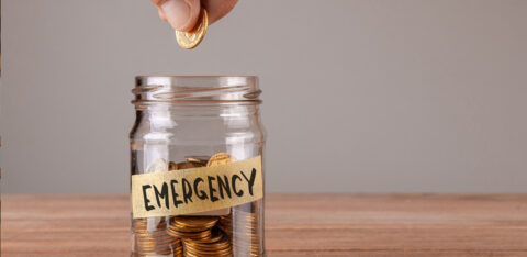 Hand placing a coin into a glass jar labeled “Emergency,” symbolizing saving money for unexpected expenses.