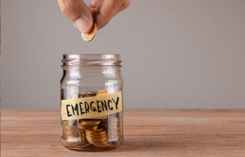 Hand placing a coin into a glass jar labeled “Emergency,” symbolizing saving money for unexpected expenses.
