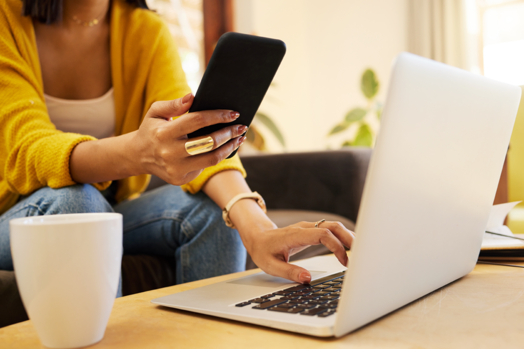 A woman multitasking with a smartphone and laptop while sitting on a couch, wearing a yellow sweater and jeans, with a coffee mug on the table.