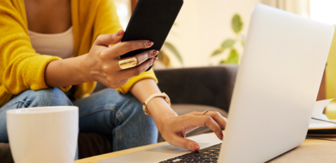 A woman multitasking with a smartphone and laptop while sitting on a couch, wearing a yellow sweater and jeans, with a coffee mug on the table.
