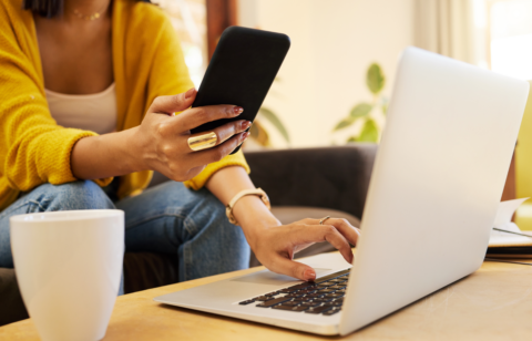 A woman multitasking with a smartphone and laptop while sitting on a couch, wearing a yellow sweater and jeans, with a coffee mug on the table.
