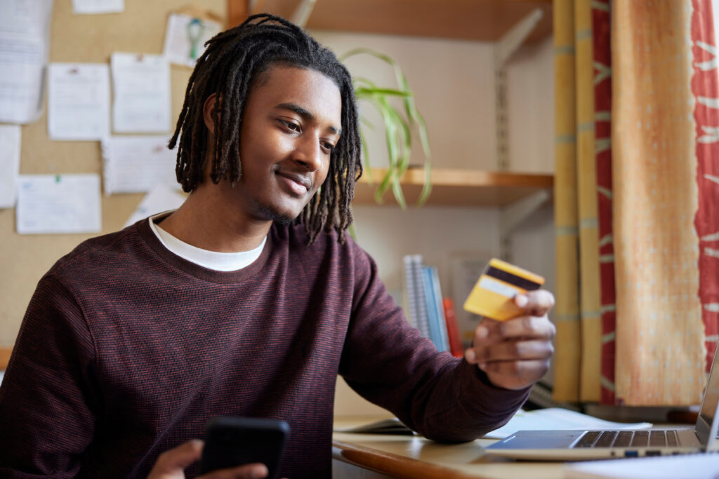 A college student holding a credit card while using a smartphone and laptop at a desk, appearing thoughtful.