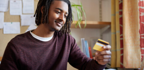 A college student holding a credit card while using a smartphone and laptop at a desk, appearing thoughtful.