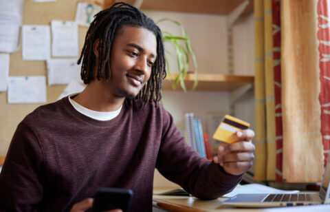 A college student holding a credit card while using a smartphone and laptop at a desk, appearing thoughtful.