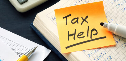 Sticky note with “Tax Help” written on it placed on a ledger book with financial figures, alongside a marker, calculator, and yellow pen.