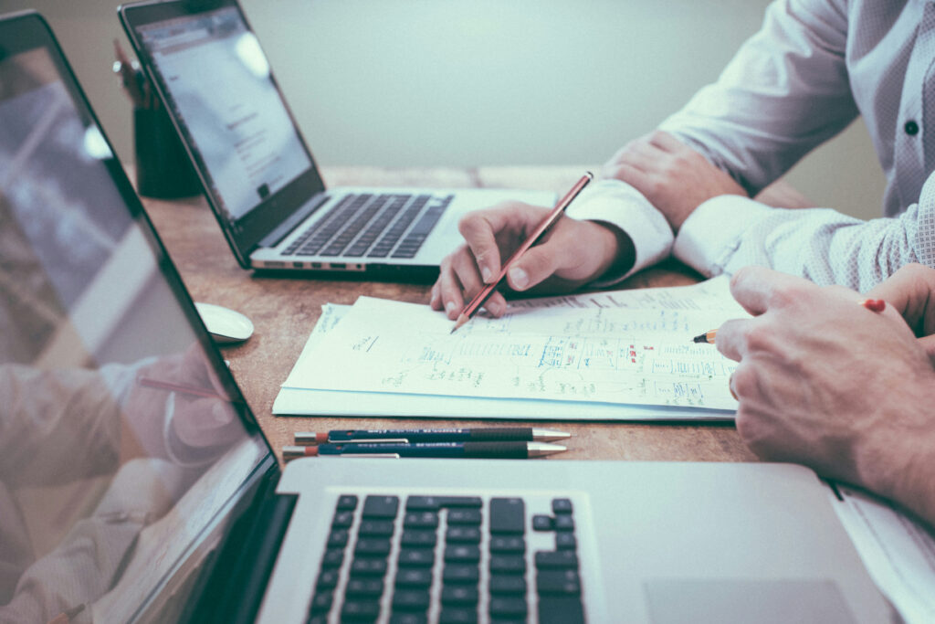Two people collaborating over documents with laptops open on a wooden desk, focusing on handwritten notes and diagrams.