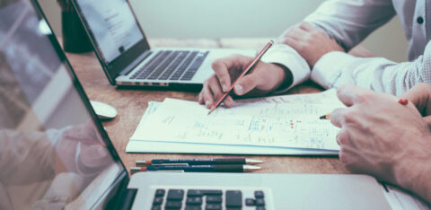 Two people collaborating over documents with laptops open on a wooden desk, focusing on handwritten notes and diagrams.