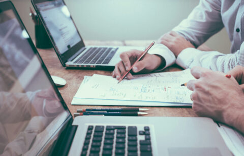 Two people collaborating over documents with laptops open on a wooden desk, focusing on handwritten notes and diagrams.