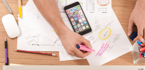 Person highlighting notes on wireframe sketches at a cluttered desk with a smartphone and colorful pens.